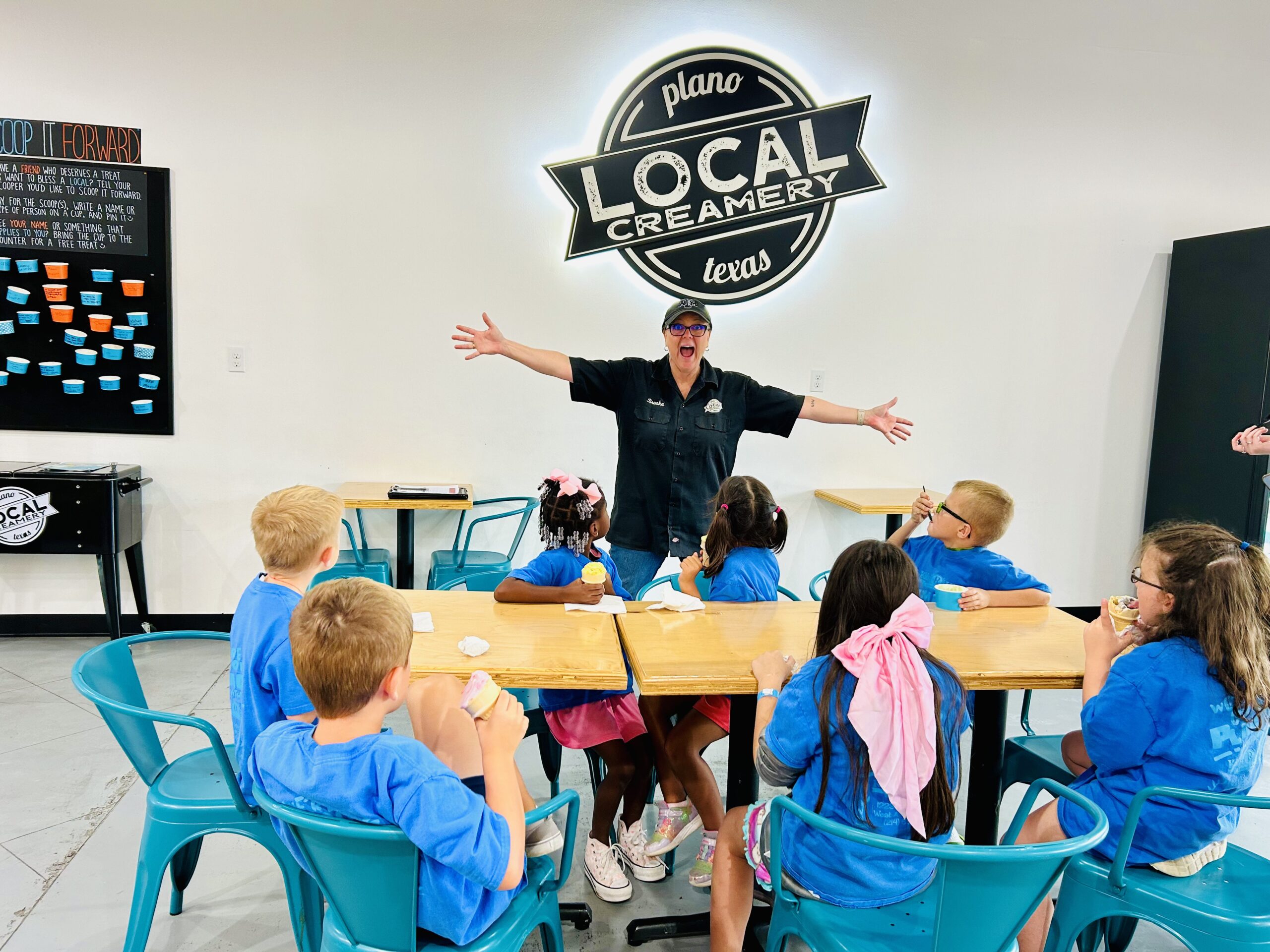 Image of Local Creamery Owner, Brooke, extending her arms with a big smile and standing in front of a group of kids eating ice cream.