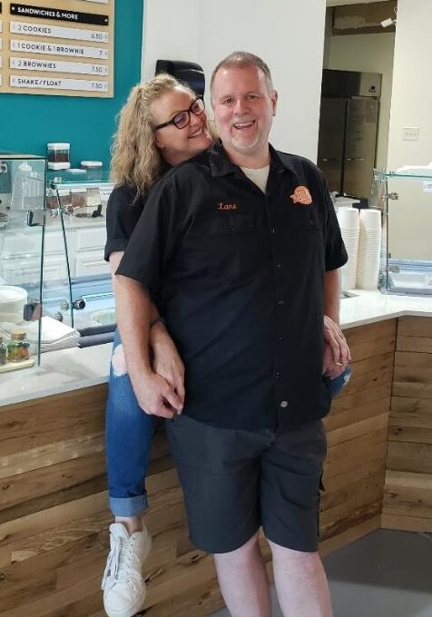 Image of Lane smiling, while Brooke sits on the counter and hugs him from behind and smiles. 