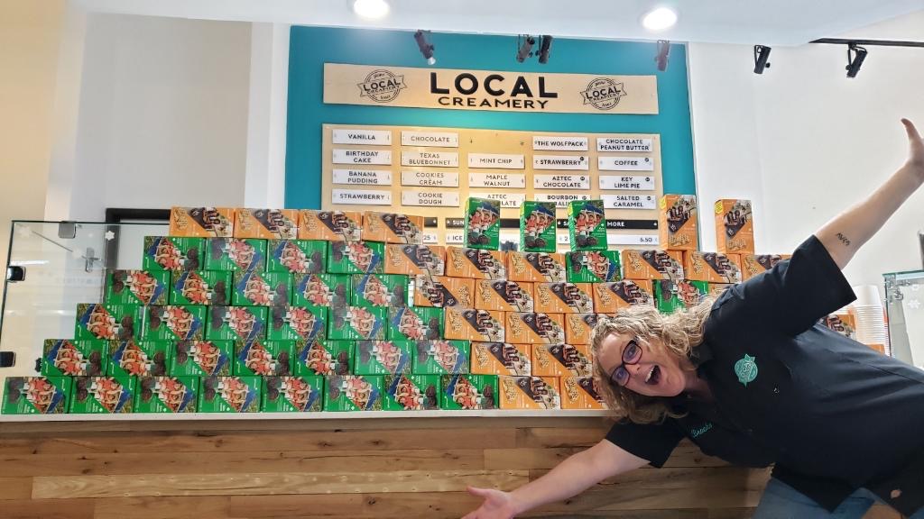 Image of a smiling Brooke showcasing stacks of Girl Scout cookie boxes at Local Creamery.